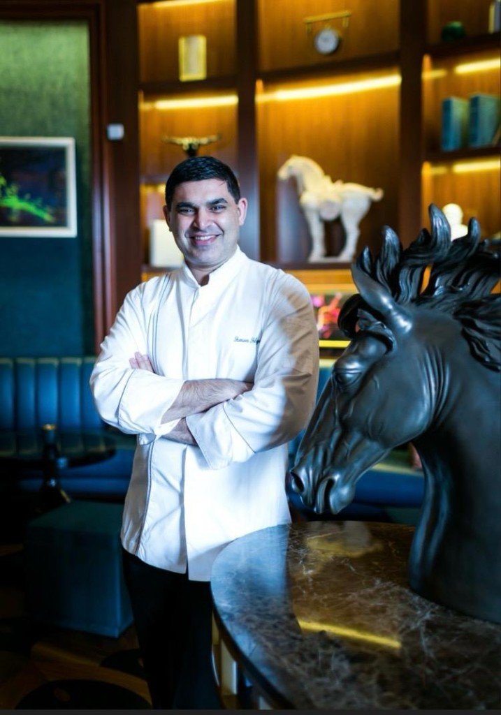 Chef Sameer Farhad Sehgal, luxury hotel chef, in a white chef coat beside a bronze horse sculpture